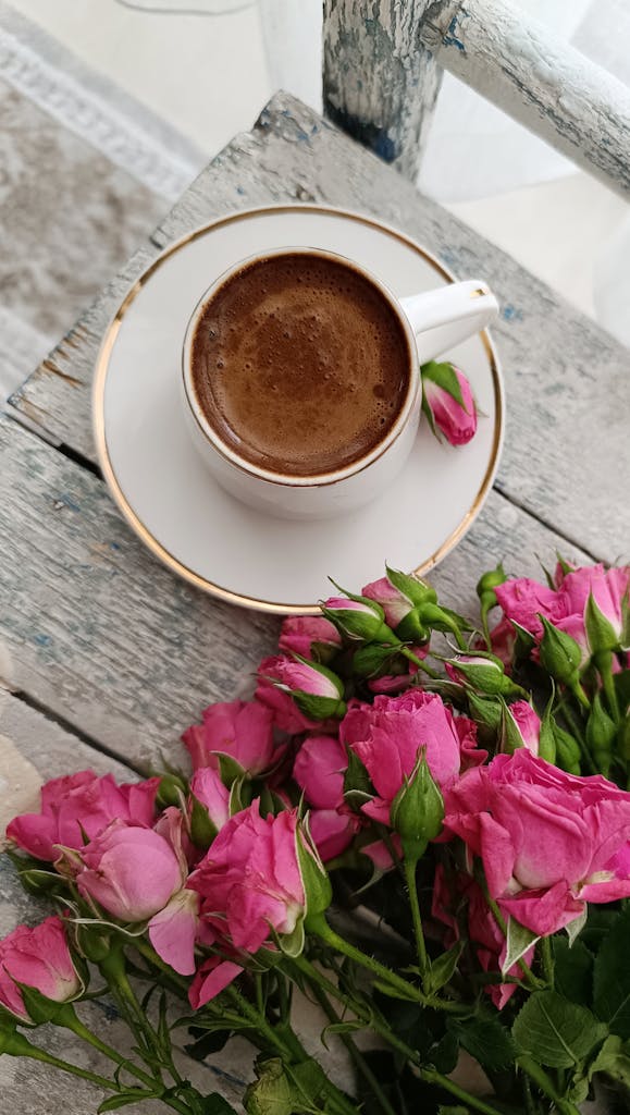A cup of coffee sits beside vibrant pink roses on a rustic wooden table, creating a cozy and inviting scene.